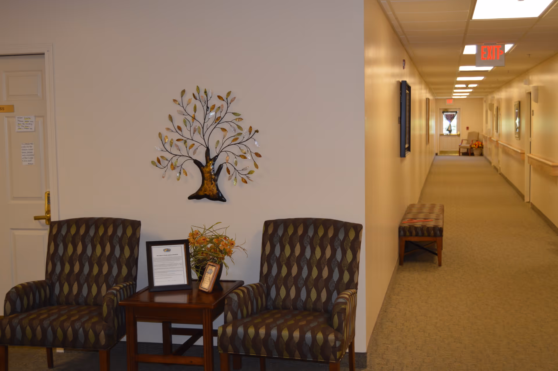 A hallway in an assisted living facility with beige walls and carpeted floor. On the left side, there are two patterned armchairs with a small wooden table between them, holding a framed document, a small picture frame, and a flower arrangement. Above the chairs, a decorative metal tree wall art is mounted. The hallway extends into the distance with benches along the right wall and an exit sign visible on the ceiling.