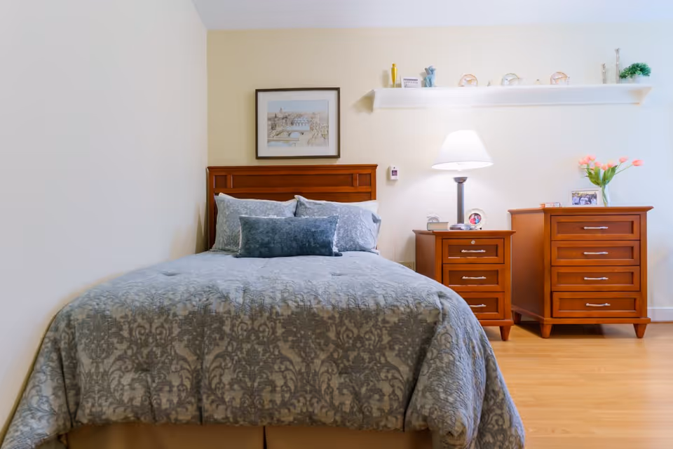 A neatly made bed with a patterned blue-gray comforter and matching pillows in a bedroom. The bed has a wooden headboard. To the right of the bed are two wooden nightstands, one with a lamp, framed photos, and a small clock, and the other with a vase of pink flowers and a framed picture. Above the nightstands is a white floating shelf with decorative items. A framed picture hangs on the wall above the headboard. The room has light-colored walls and wooden flooring.