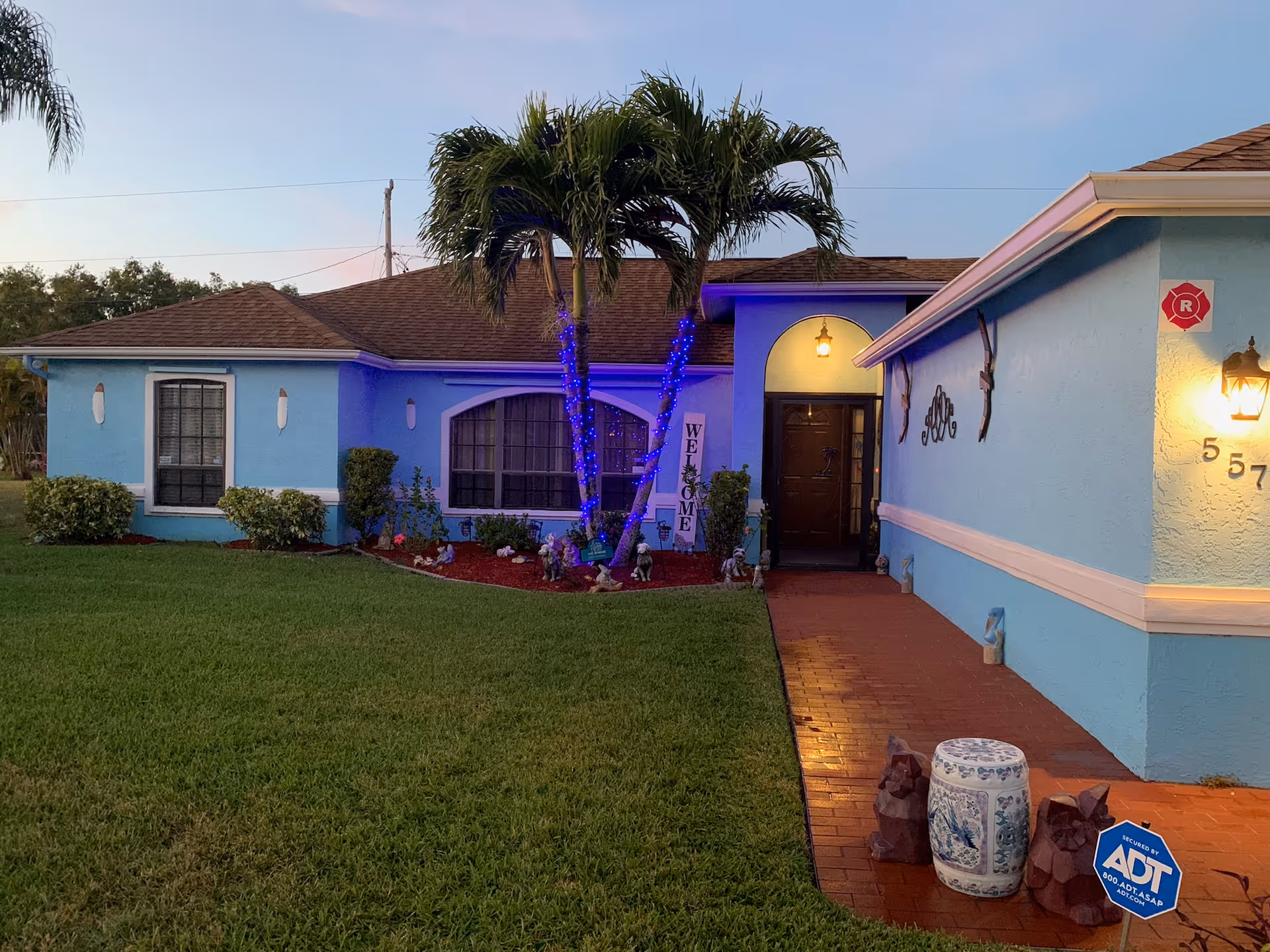 Exterior view of a single-story light blue house with a brown shingle roof during dusk. There is a palm tree wrapped with blue lights in front of the house, a welcome sign near the entrance, and various small garden decorations. The house has a well-maintained green lawn and a red brick walkway leading to a dark brown front door. A lit lantern is mounted on the wall near the house number 557, and a blue ADT security sign is placed in the garden near the walkway.
