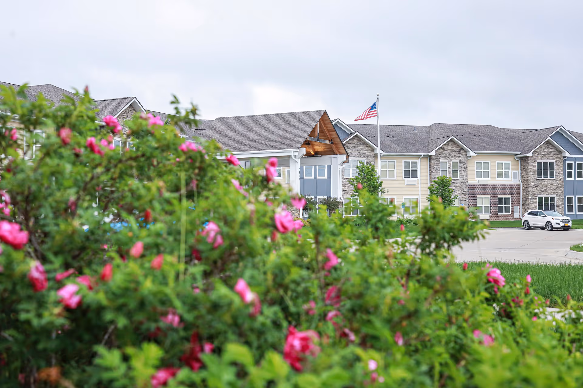 Front exterior of a senior living building with flowering bushes in the foreground and an American flag flying near the entrance.