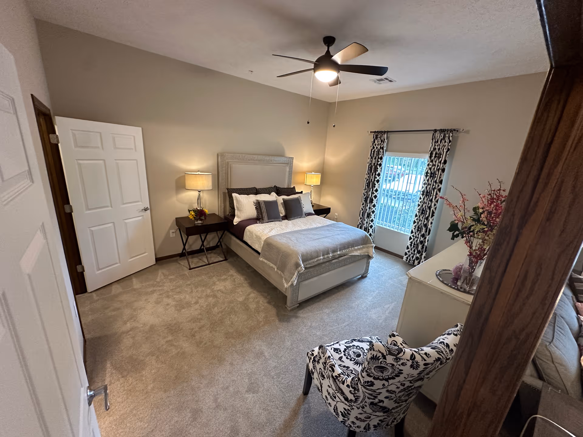 A cozy bedroom with a bed centered against the wall, flanked by two nightstands each with a lamp. The bed is made with white and dark pillows and a light-colored blanket. There is a patterned armchair in the foreground, a dresser with a floral arrangement on top, a window with patterned curtains, and a ceiling fan with a light.