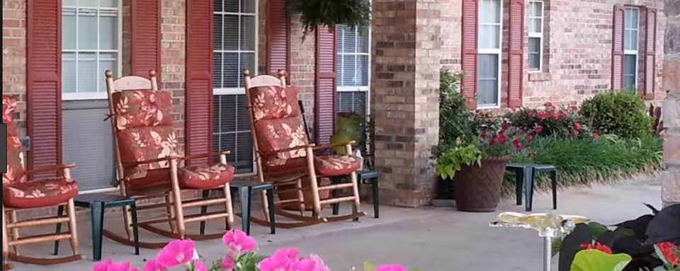 Front porch of a brick senior living facility with floral-cushioned rocking chairs, potted plants, and blooming flowers in the foreground.
