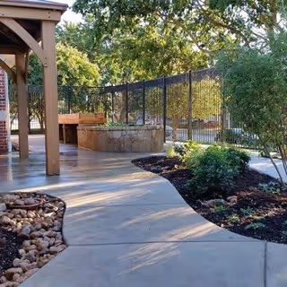 Outdoor patio area at a senior living facility featuring a curved concrete walkway, landscaped garden beds with shrubs and rocks, a wooden pergola structure, and a stone fire pit surrounded by benches. Trees and a black metal fence are visible in the background.