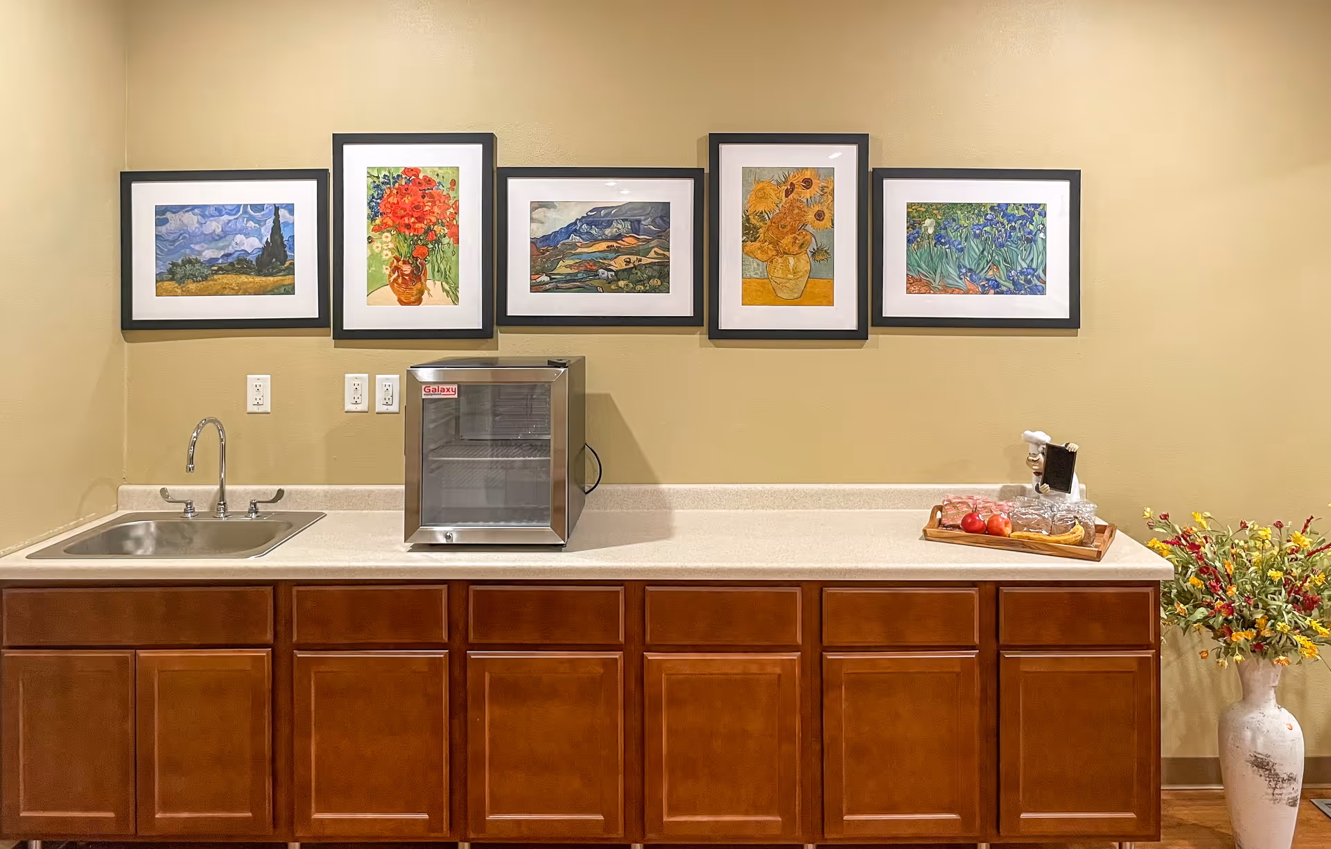 A kitchenette area with a stainless steel sink on the left, a small glass-front refrigerator in the center, and a countertop with a tray of snacks and a vase of flowers on the right. Above the counter are five framed paintings of colorful floral and landscape scenes. The cabinets below the counter are wooden with a warm brown finish.