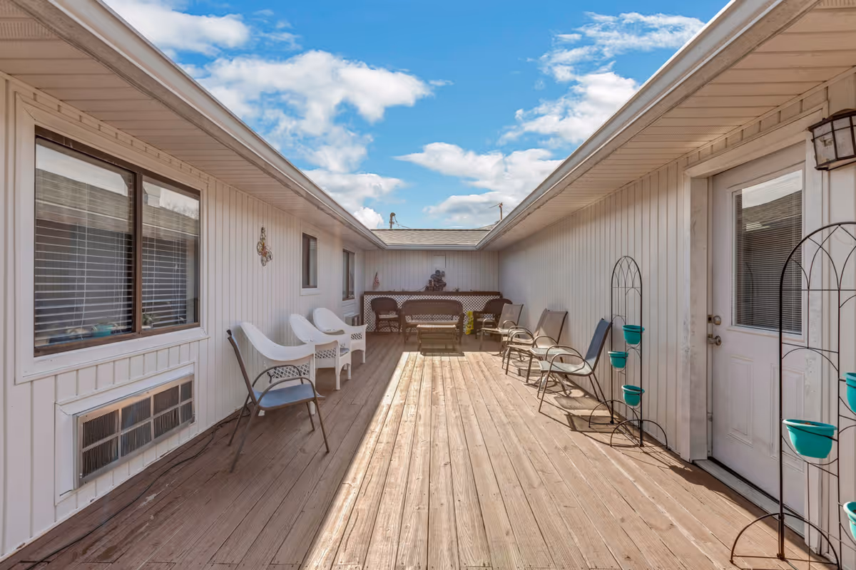 Outdoor wooden deck area between two building sections with several chairs lined up along the walls, a small table and bench at the far end, white siding on the buildings, and a partly cloudy blue sky above.