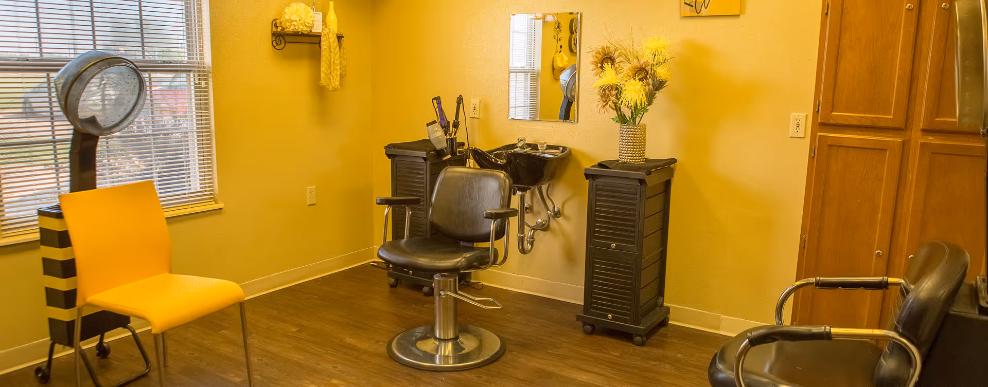 Interior view of a small salon area with a black salon chair in front of a wall-mounted mirror and a hair washing sink. There is a yellow chair near a window with blinds, a black rolling cart with hair tools, a cabinet with a vase of yellow flowers, and wooden cabinets on the right side.
