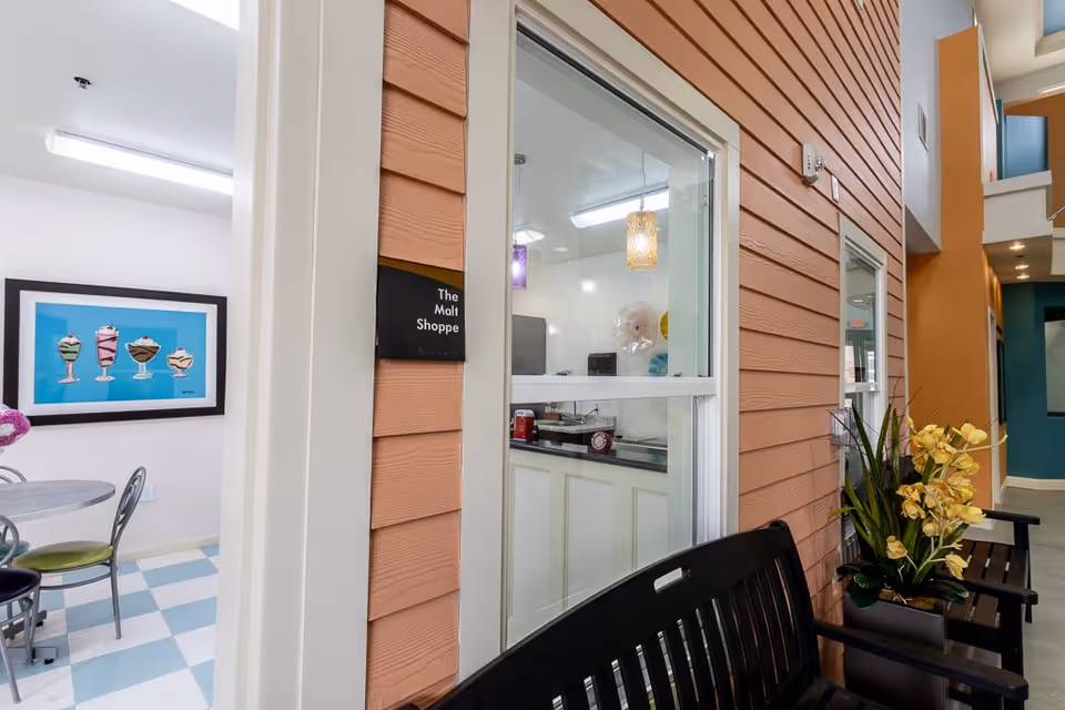 Interior view of a senior living facility showing a small dining area with a round table and chairs, a colorful framed picture of ice cream sundaes on the wall, and a window into a room labeled 'The Malt Shoppe'. The hallway features orange siding walls, a black bench with yellow flowers in a planter, and a glimpse of other rooms and corridors.