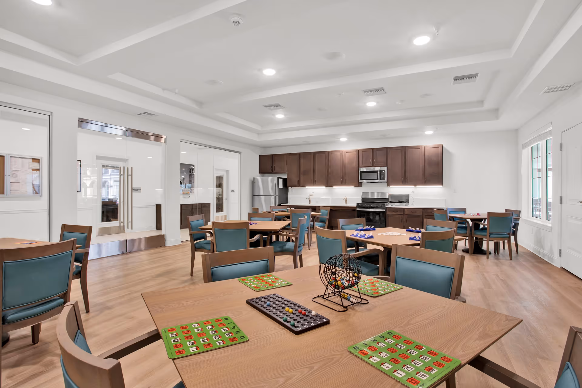 A bright community room with multiple wooden tables and teal cushioned chairs arranged for a bingo game. The tables have bingo cards and a bingo cage with balls. The room features a kitchen area with dark wood cabinets, a stainless steel refrigerator, microwave, and stove. The floor is light wood, and the ceiling has recessed lighting. Glass double doors and windows provide additional light.