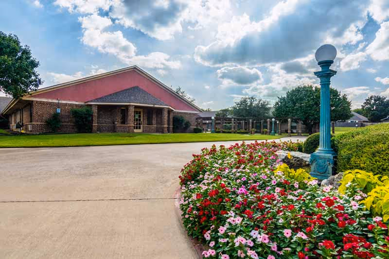Exterior view of a healthcare facility building with a brick and pink facade, surrounded by a well-maintained garden featuring colorful flowers and a blue lamp post under a partly cloudy sky.
