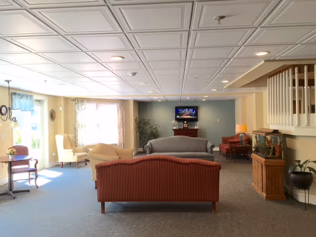 A spacious living room area with multiple upholstered sofas and armchairs arranged facing a wall-mounted television. The room has a light blue carpet, a white coffered ceiling, and large windows letting in natural light. There is a wooden cabinet with a fish tank on the right side and a small table with a flower vase on the left. The walls are painted in soft yellow and blue tones, and there are some plants and lamps adding to the cozy atmosphere.
