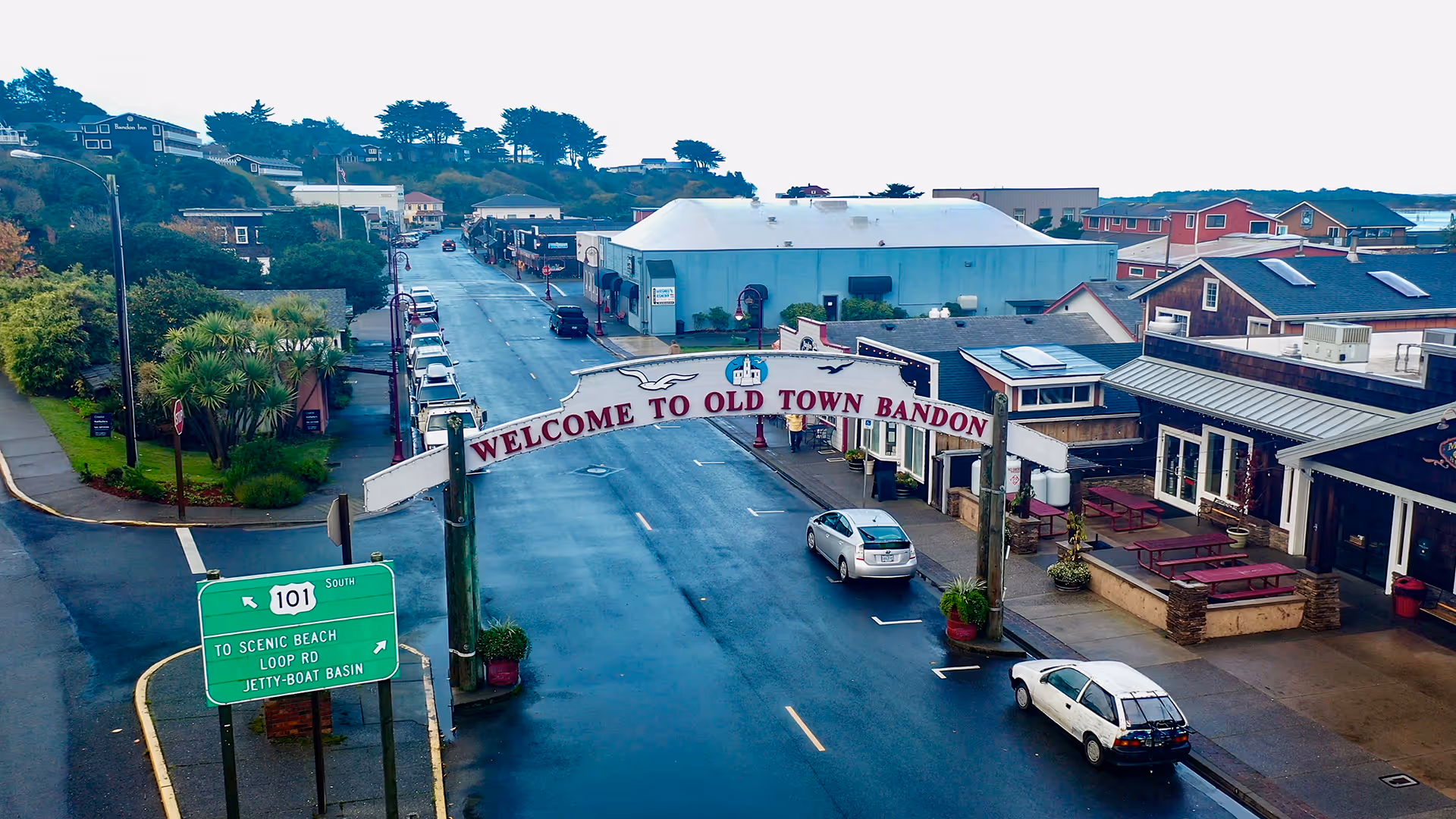 Street-level view of Old Town Bandon with an arch reading 'WELCOME TO OLD TOWN BANDON' over a wet road lined with shops and parked cars.