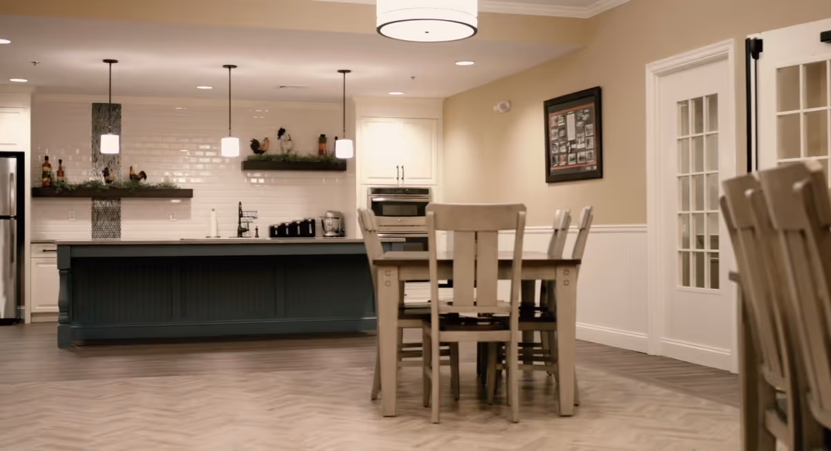 A modern kitchen and dining area in a senior living facility featuring a large kitchen island with pendant lights above, white cabinetry, stainless steel refrigerator, built-in oven, and a wooden dining table with chairs. The floor has a herringbone pattern, and there is a framed picture on the beige wall.