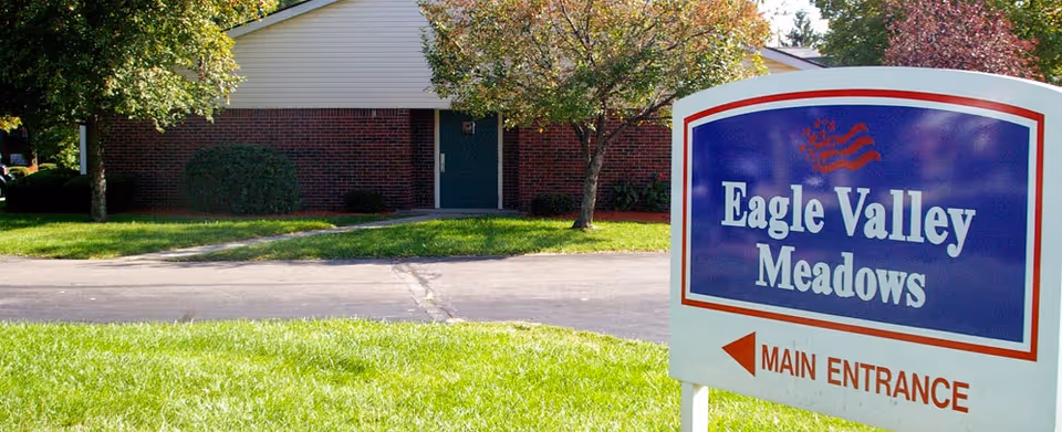 Outdoor view of Eagle Valley Meadows facility showing a sign with the facility name and an arrow pointing to the main entrance. The background includes a brick building, trees, and green grass.