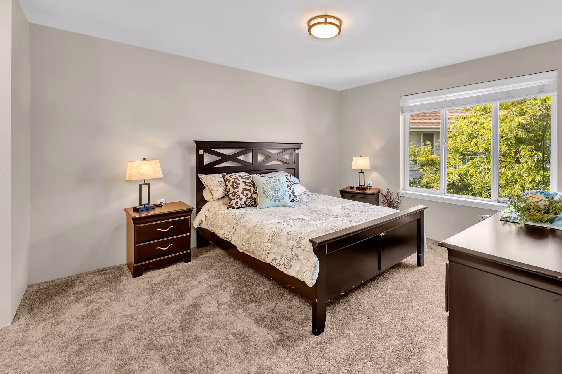 Bright furnished bedroom featuring a dark wood bed, matching nightstands and dresser, carpeted floor, and a large window with a view of trees.