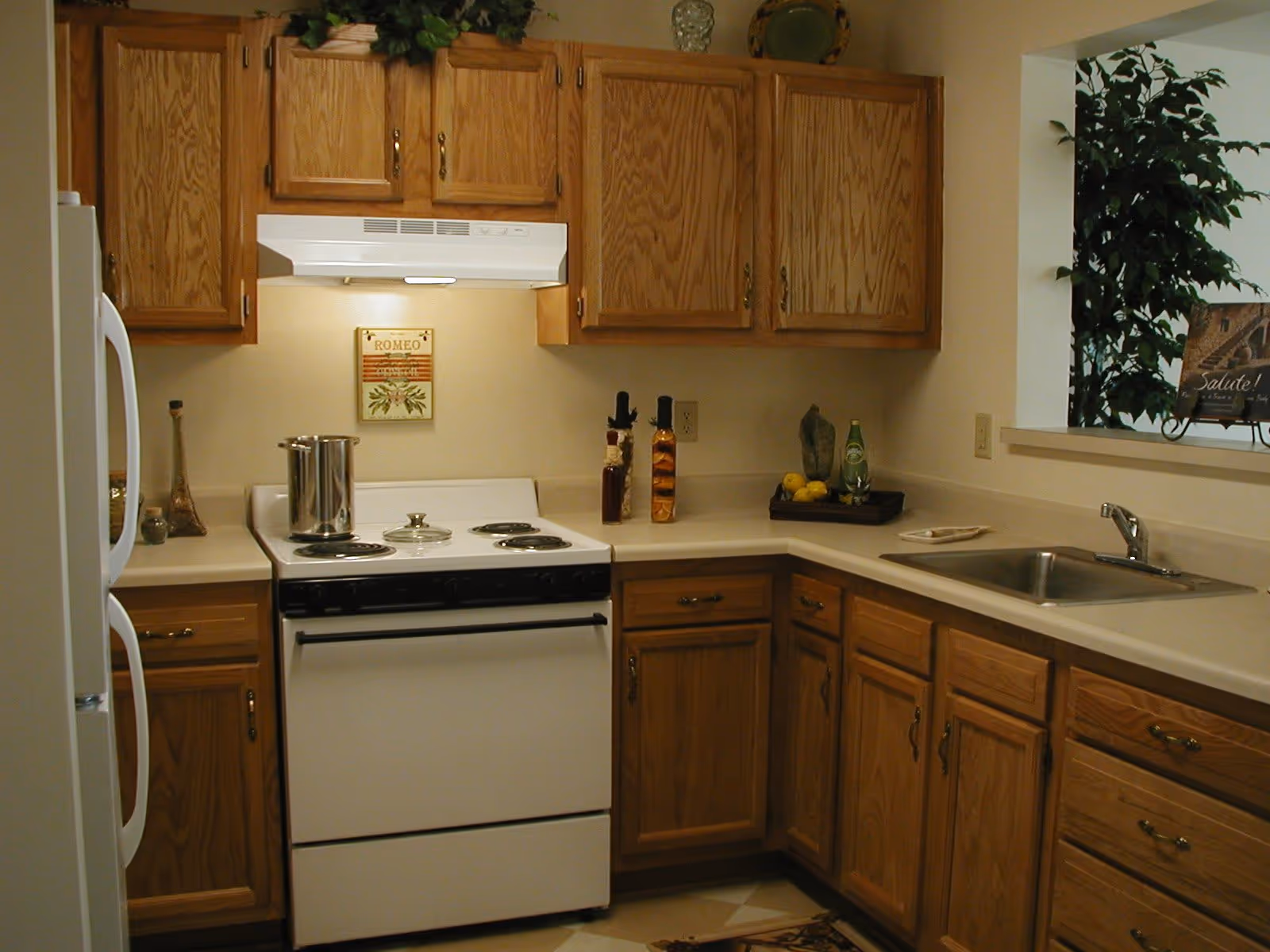 A kitchen with wooden cabinets, a white electric stove with a pot on it, a white refrigerator, a stainless steel sink, and various decorative items on the countertops including bottles, a tray with lemons, and a small framed sign near a window with a plant visible outside.