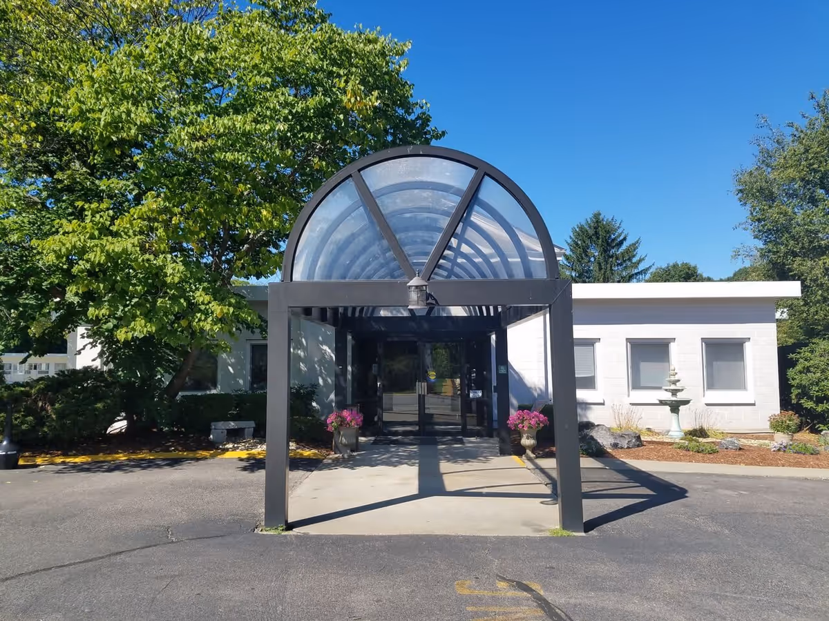 Entrance to Cedar View Rehabilitation & Healthcare Center featuring a covered walkway with a black metal frame and transparent curved roof. There are two flower pots with pink flowers on either side of the entrance. The building is white with several windows, and there are trees and landscaping around the entrance under a clear blue sky.