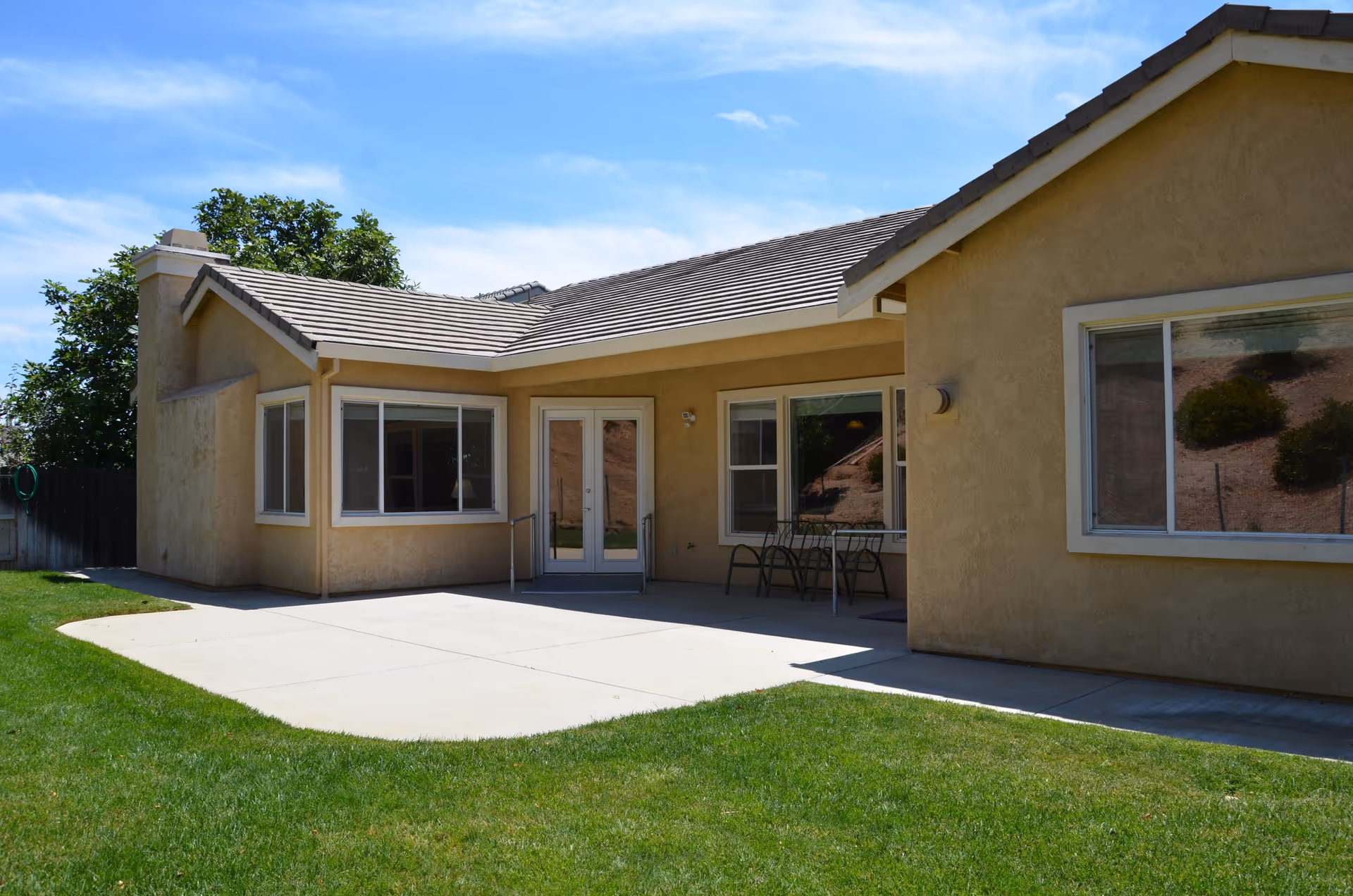 Exterior view of a single-story beige stucco building with a tiled roof, large windows, a glass double door, and a small patio area with a table and chairs. The building is surrounded by a green lawn and a clear blue sky is visible above.
