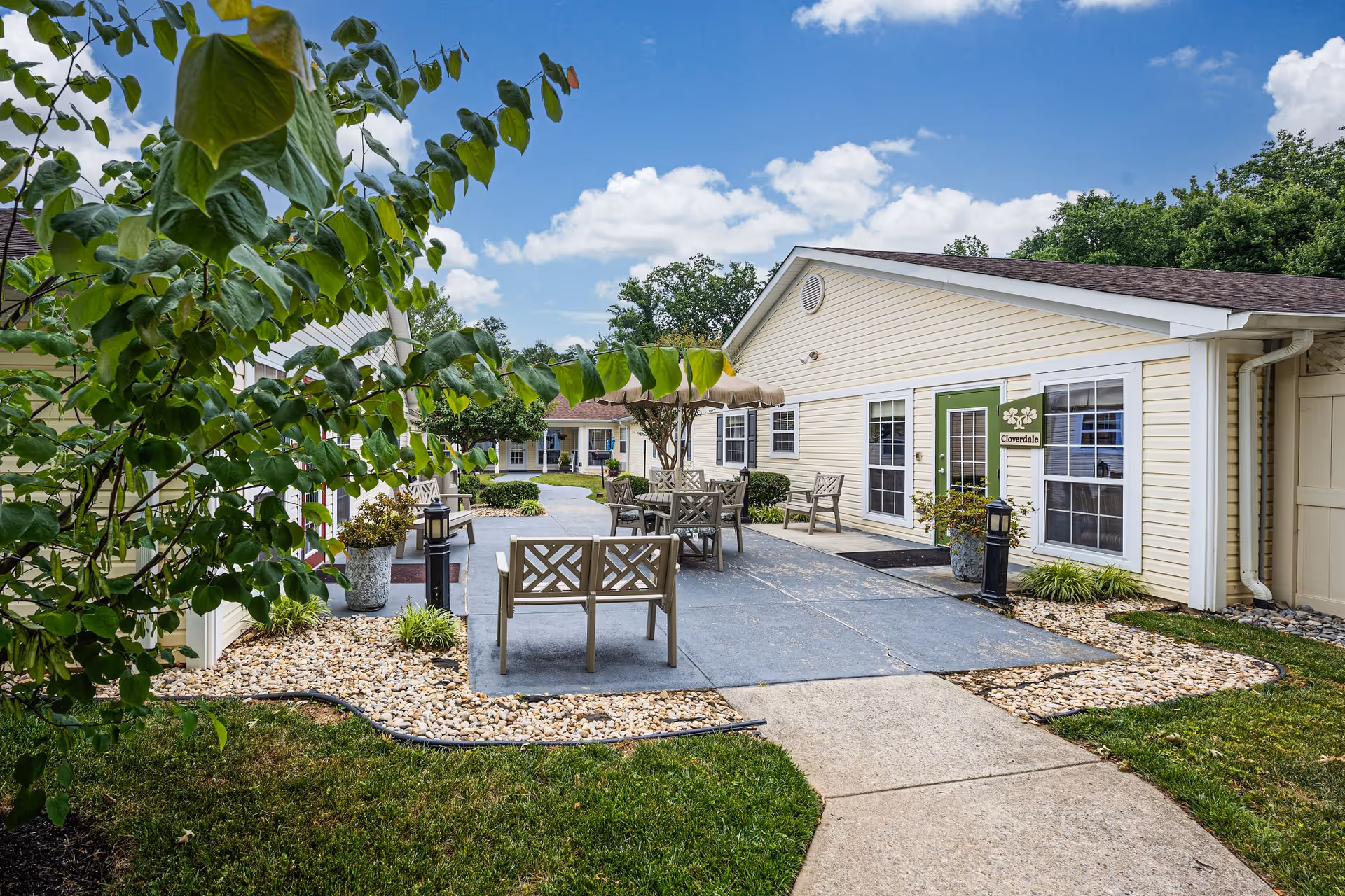 Outdoor patio area at Arden Courts - ProMedica Memory Care Community in Silver Spring, featuring beige buildings with white trim, several wooden benches and tables with umbrellas, landscaped with green grass, bushes, and trees under a blue sky with scattered clouds.