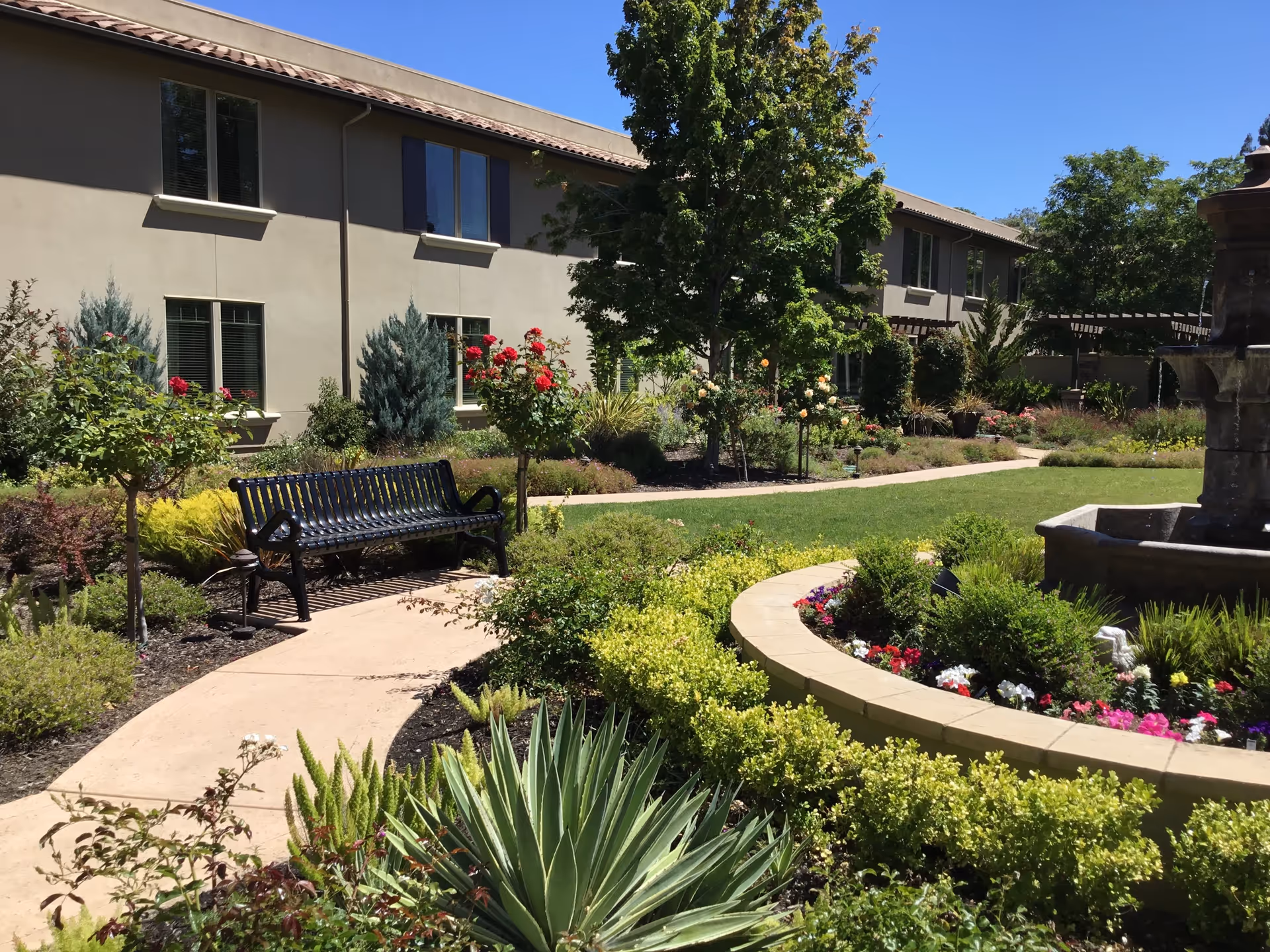 A landscaped outdoor garden area at Oakmont of Concord featuring a curved concrete pathway, a black metal bench, various green shrubs and flowering plants, a large tree, and a stone fountain surrounded by colorful flowers. The background shows a beige two-story building with multiple windows under a clear blue sky.