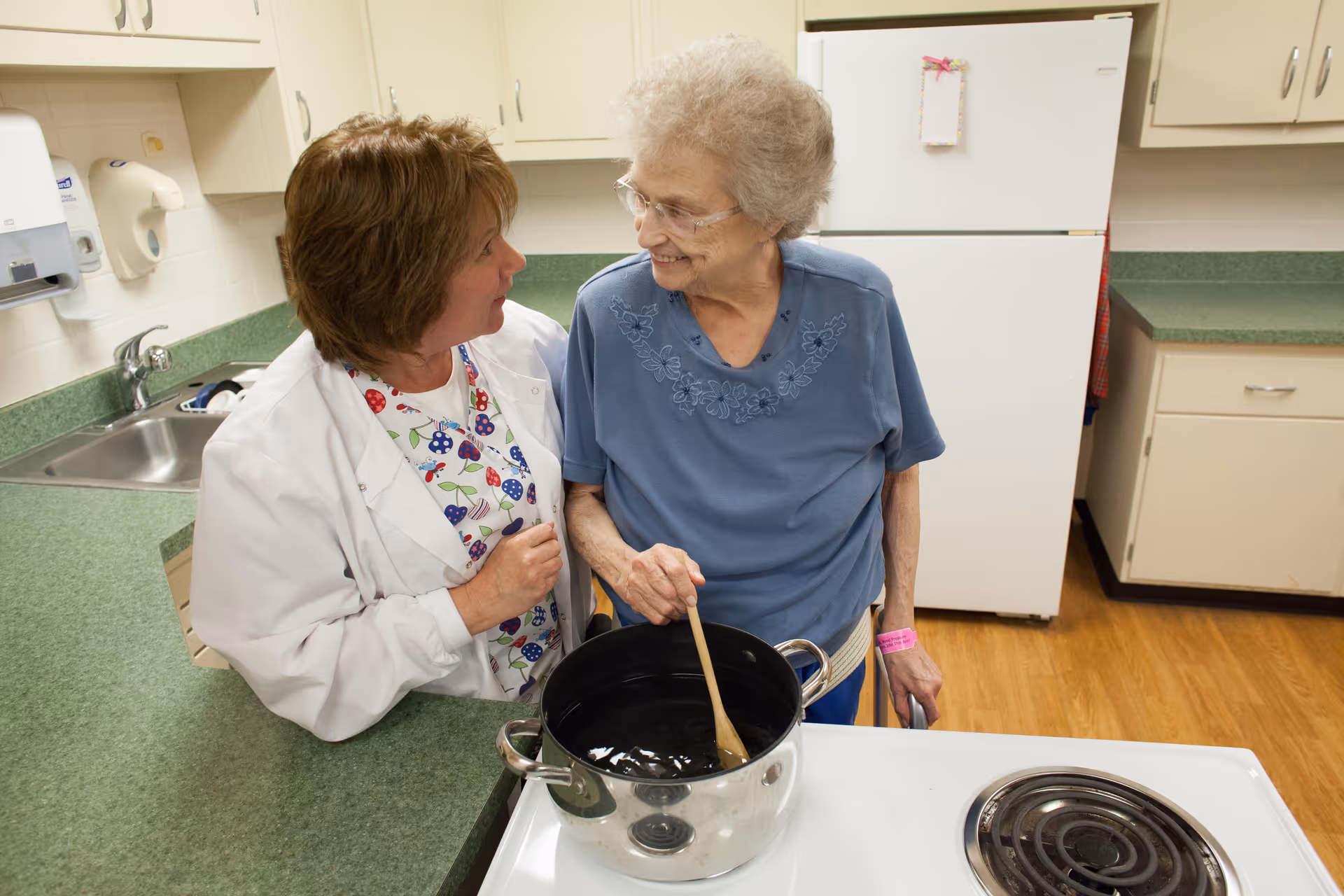 An elderly woman stirring a pot on a stove in a kitchen, smiling and looking at a healthcare worker who is leaning on the counter and engaging with her. The kitchen has green countertops, white cabinets, a sink, and a white refrigerator in the background.