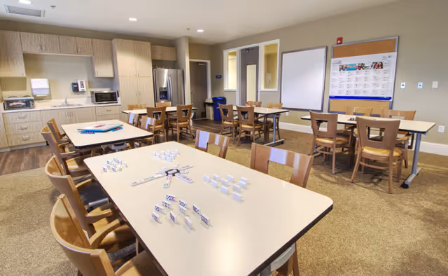 A bright and spacious activity room in Carmel Village featuring multiple tables and wooden chairs arranged for group activities. One table in the foreground has dominoes set up for a game. The room includes a kitchenette area with cabinets, a microwave, and a refrigerator. Bulletin boards and a whiteboard are mounted on the wall.