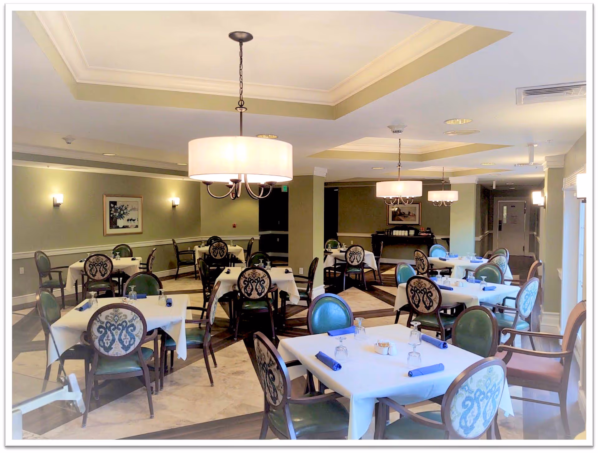 A dining room with multiple tables covered in white tablecloths, each set with blue napkins and upside-down glasses. The chairs have green seats and patterned backs. The room is softly lit with hanging chandeliers and wall sconces, and there are framed pictures on the walls.