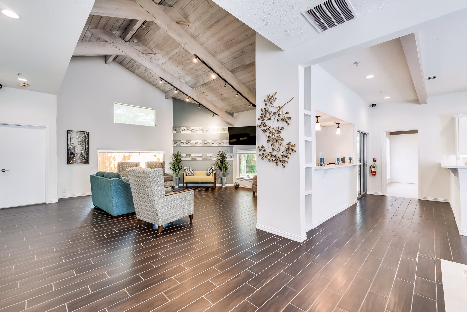 Bright open common room with vaulted wooden ceiling, seating area of chairs and a sofa, decorative wall art, and dark tile floor.