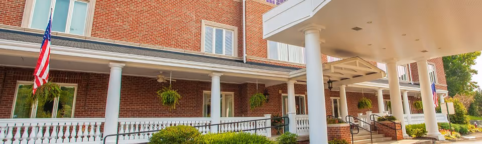 Exterior view of a brick senior living facility with white columns supporting a covered entrance. There are hanging plants along the porch, an American flag, and well-maintained bushes and greenery in front of the building.