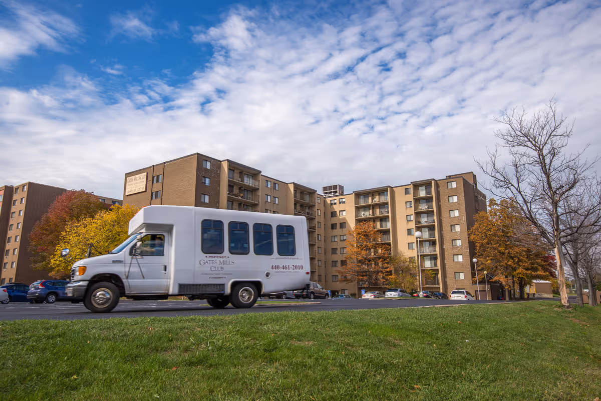 A white Gates Mills Club shuttle van parked in front of a multi-story brick senior living building under a partly cloudy sky.