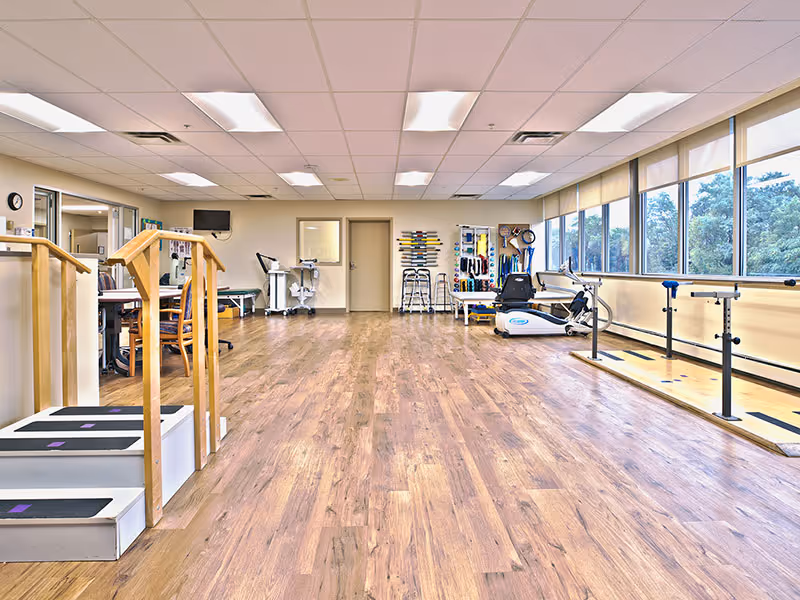A bright physical therapy/exercise room with parallel bars, steps, equipment, and large windows along one wall.