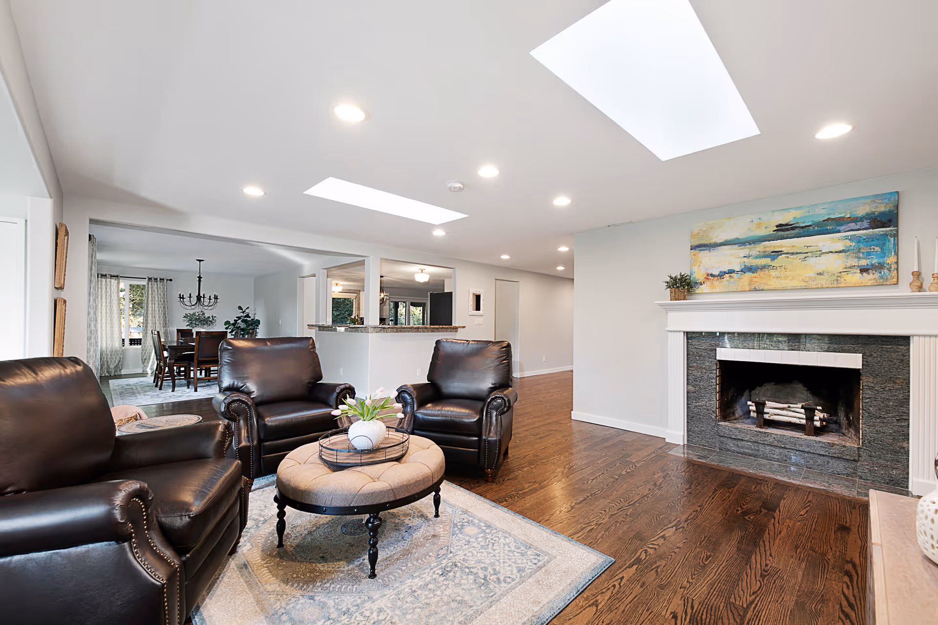 A bright living room with three dark leather armchairs around a round ottoman on a rug, hardwood floors, skylights, and a fireplace.