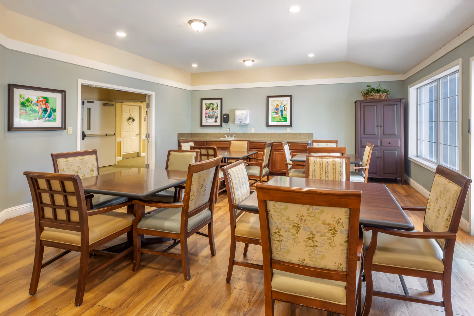 A bright dining room in a senior living facility with several wooden tables and cushioned chairs arranged neatly. The room has light green walls, wooden flooring, and large windows letting in natural light. There are framed pictures on the walls, a cabinet with a plant on top, and a counter with a sink and paper towel dispenser in the background.