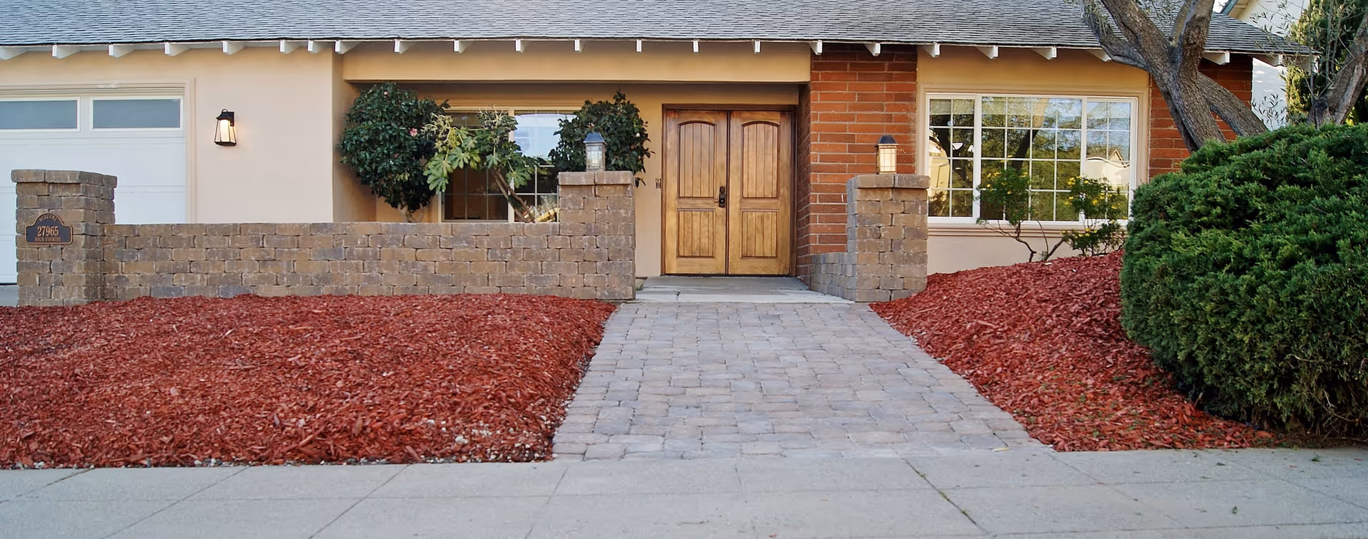 Front exterior view of a single-story house with a brick and stucco facade, a double wooden front door, a large window, a paved walkway leading to the entrance, and landscaped areas with red mulch and green bushes.