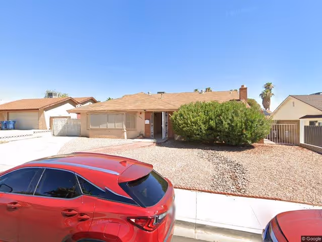 Single-story residential house with a brown roof and beige exterior walls, featuring a large bush in the front yard covered with gravel. A red car is parked on the street in front of the house. The sky is clear and blue.