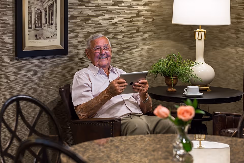 An elderly man sitting in a leather armchair in a cozy room, smiling and holding a tablet. Behind him is a round table with a white lamp, a potted plant, and a cup and saucer. A framed black and white picture hangs on the textured wall.