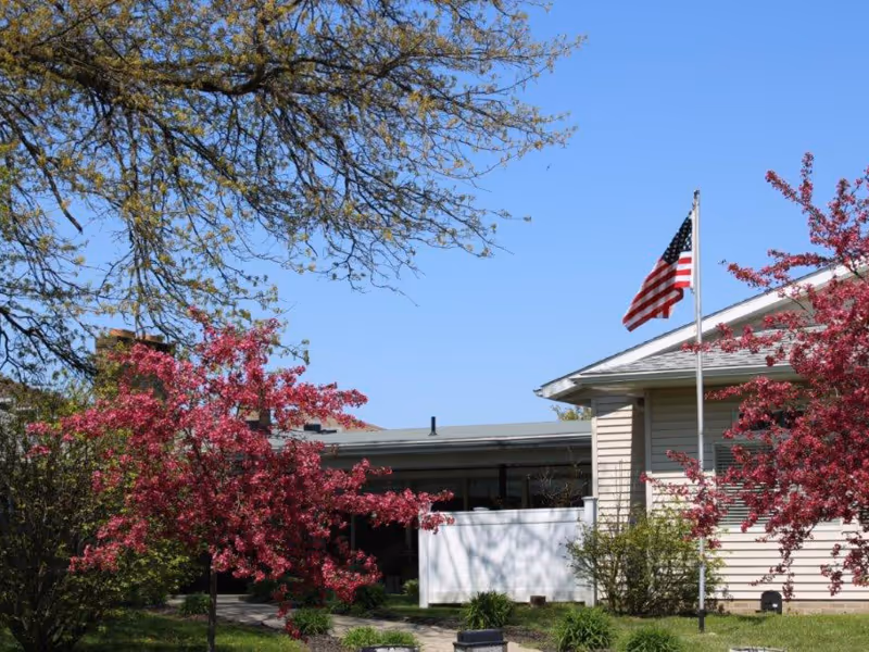 Exterior view of a single-story building with an American flag and pink-flowering trees in front.