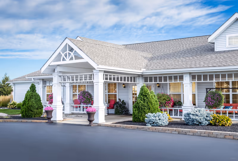 Exterior view of a single-story building with a covered entrance supported by white columns. The building has light gray siding and a shingled roof. There are decorative plants and flowers in pots and landscaped beds around the entrance. Red chairs are visible on the porch area under the covered entrance. The sky is partly cloudy and blue.
