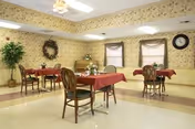 A dining room with several tables covered in red tablecloths, each surrounded by wooden chairs. The walls are decorated with floral wallpaper, a wreath, and a clock. There are two windows with curtains and a potted plant in the corner.