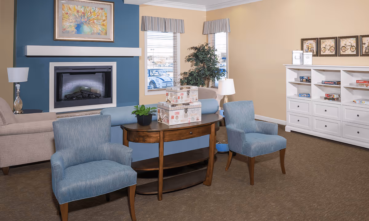 Common room with blue armchairs and a sofa facing a fireplace, a wooden console table, and white shelving with games.
