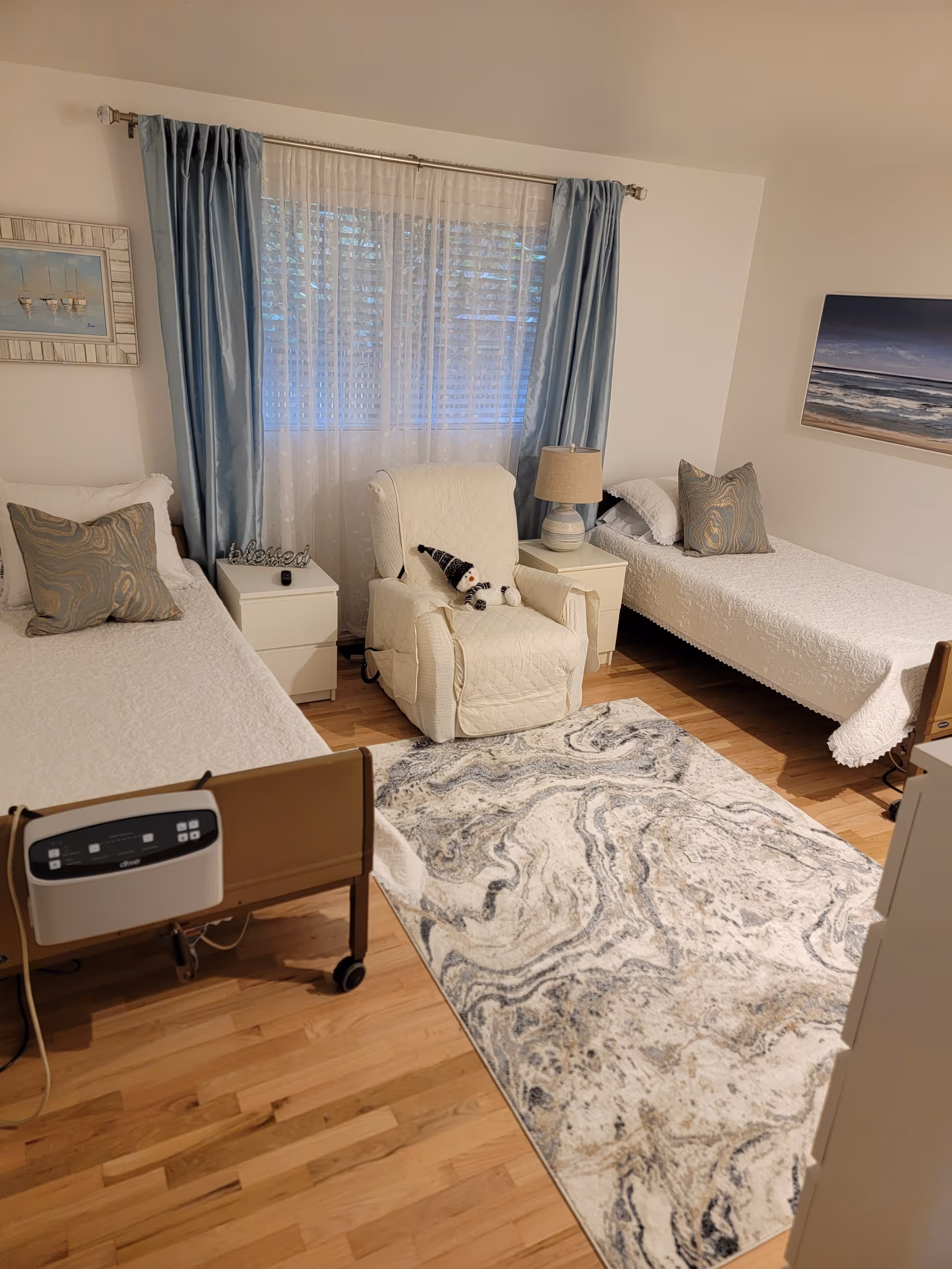 Shared bedroom with two single beds, a recliner between them, nightstands and lamp by a curtained window, and a patterned rug on hardwood floors.