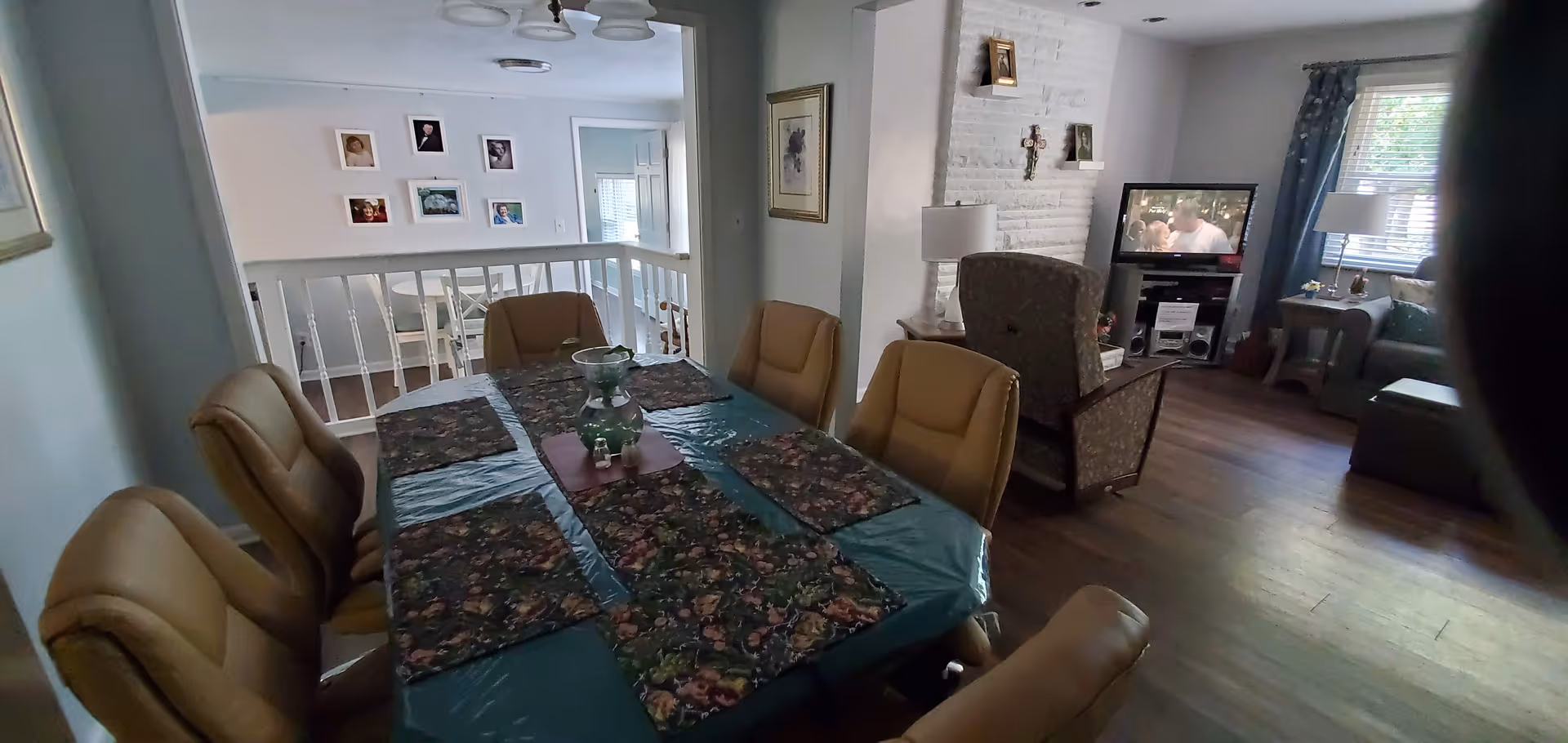 Interior view of a senior living facility showing a dining area with a table covered by a blue tablecloth and floral placemats surrounded by six tan chairs. Adjacent to the dining area is a living room with a patterned armchair, a TV on a stand, a side table with a lamp, and a window with blue curtains letting in natural light. The walls are decorated with framed pictures and a cross above the fireplace.