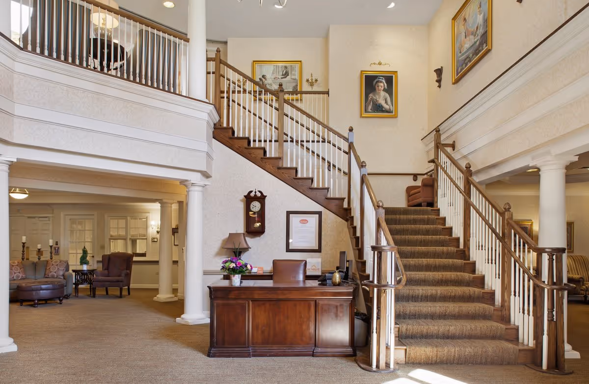 Interior view of a senior living facility lobby with a wooden reception desk, a staircase with wooden handrails and carpeted steps leading to the upper floor, and framed paintings on the walls. There are columns supporting the upper floor balcony, and seating areas with chairs and sofas are visible in the background.