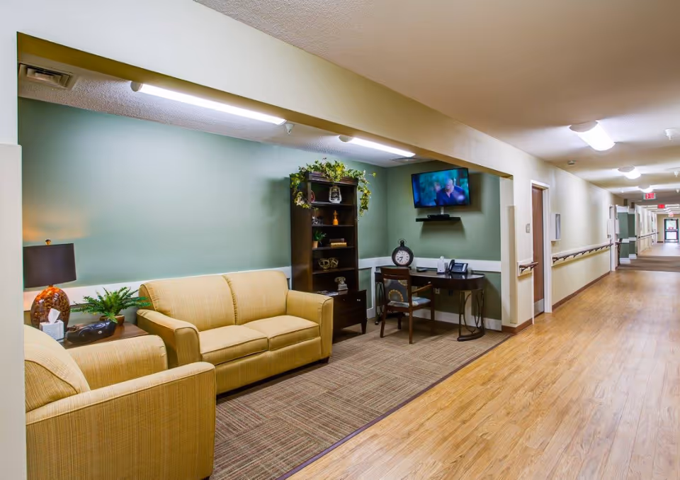 A cozy seating area in a senior living facility hallway featuring a beige couch and armchair, a wooden bookshelf with decorative items, a desk with a chair, a clock, and a wall-mounted TV. The hallway has wooden flooring, handrails, and doors leading to rooms.