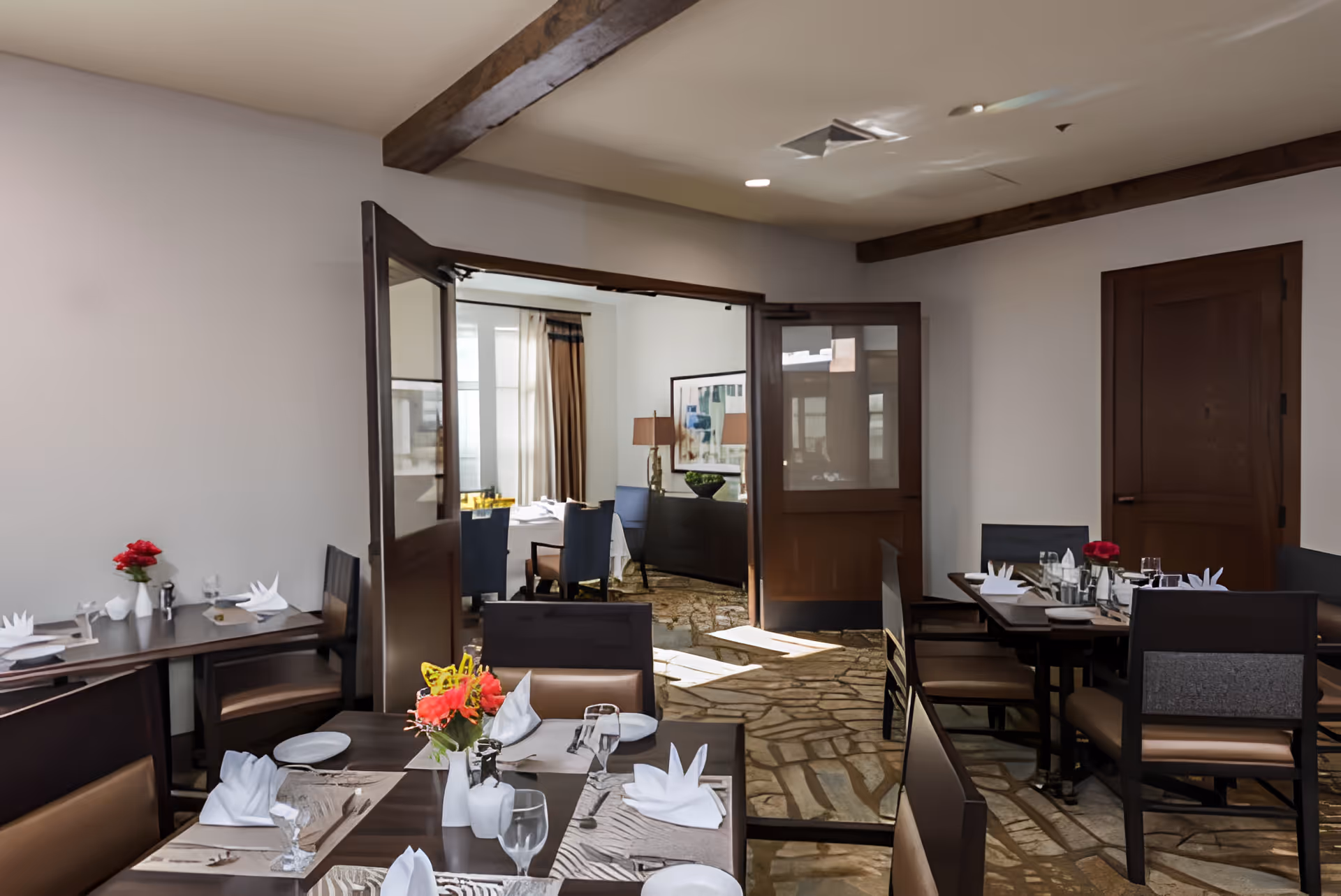 Interior view of a dining room in a senior living facility with several tables set with white napkins, plates, glasses, and silverware. The room features wooden beams on the ceiling, stone flooring, and an open doorway leading to another room with additional seating and a lamp on a sideboard.