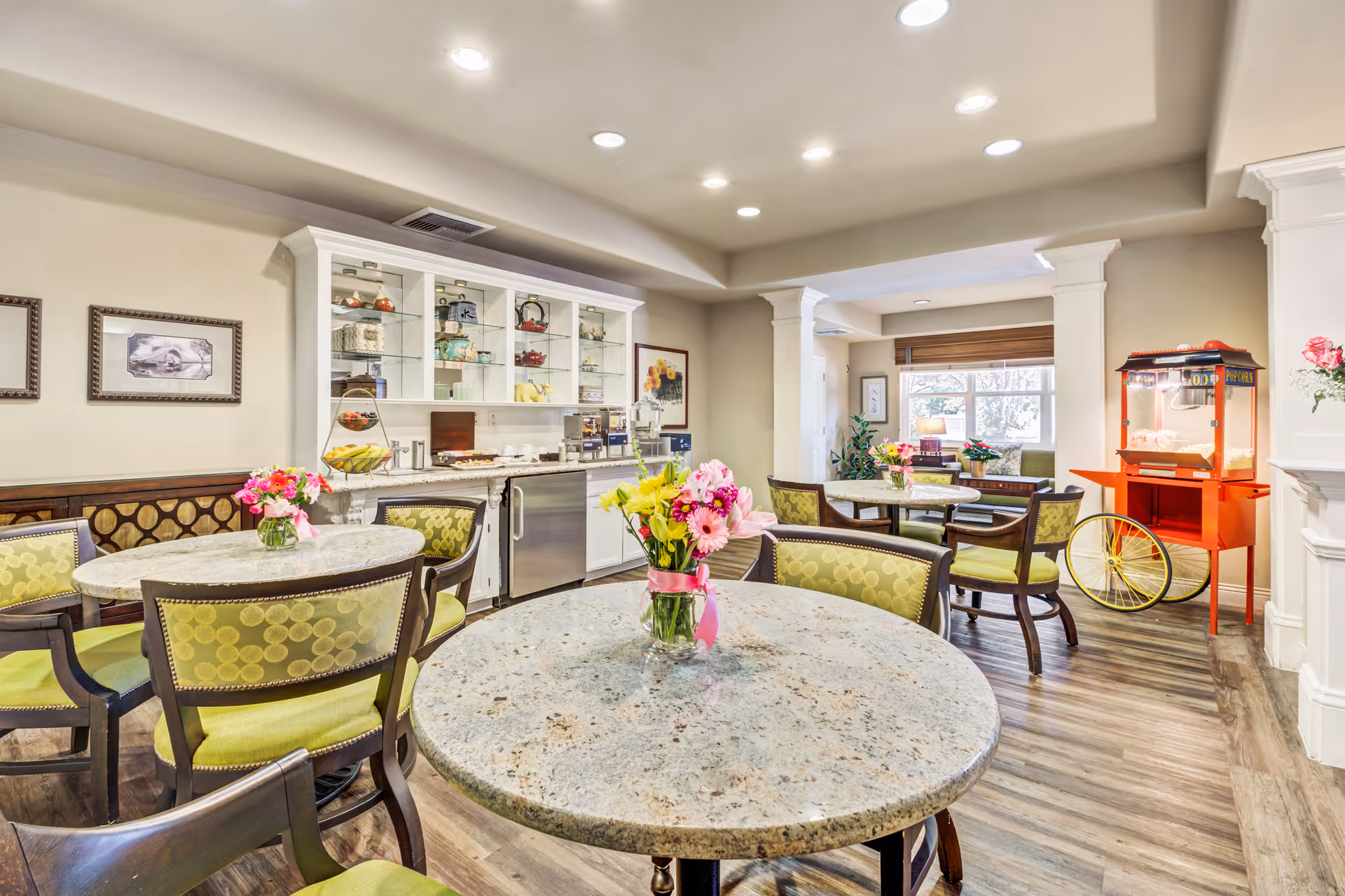 A bright and inviting dining area with round tables topped with flower vases, green cushioned chairs, a popcorn machine on the right, and a white cabinet with glass shelves displaying teapots and other items. The room has wood flooring, recessed lighting, and a window letting in natural light.