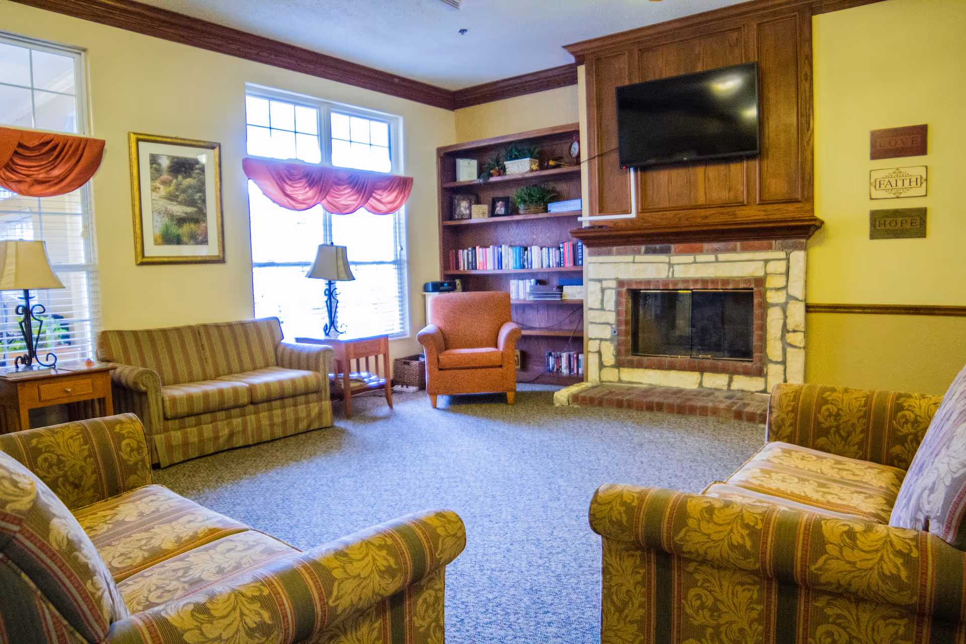 Cozy sitting room with patterned sofas and armchairs arranged around a stone fireplace and wall-mounted TV, bookshelves and a bright window.