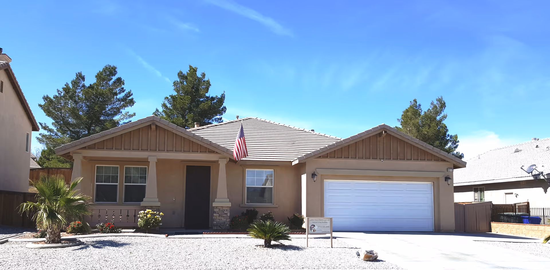 Single-story beige house with a tiled roof, an American flag hanging near the front door, a two-car garage, and a gravel front yard with small palm trees and bushes under a clear blue sky.