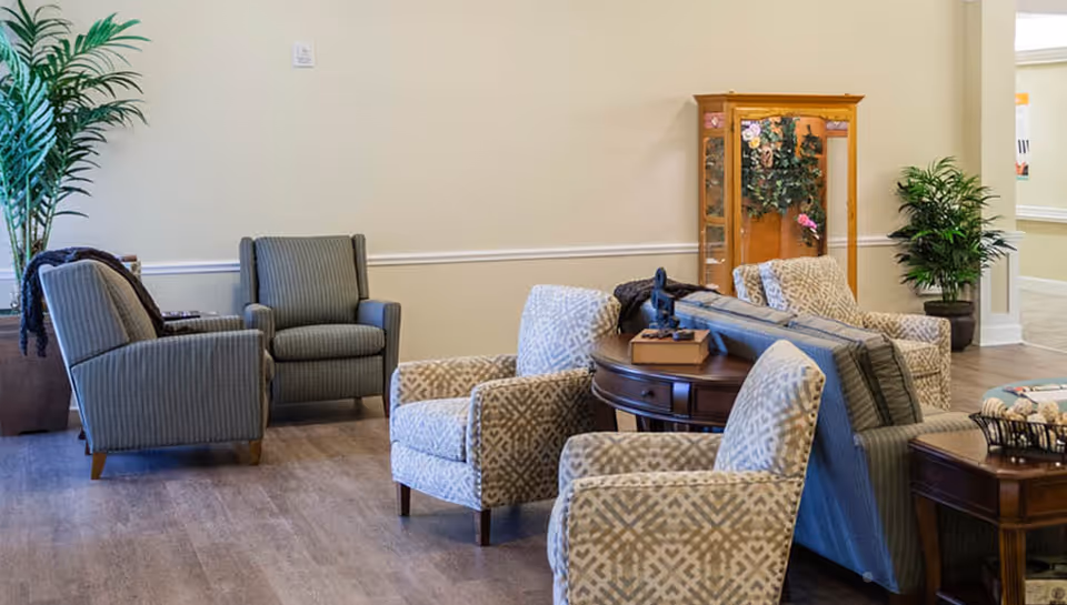 A bright common room with patterned armchairs, a sofa, side tables, and potted plants.
