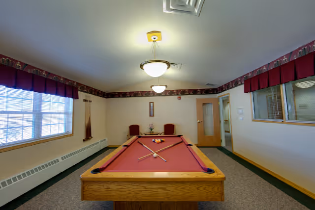 A recreation room with a red felt pool table in the center, two pool cues resting on the table, and a rack of billiard balls. The room has beige walls with a decorative border near the ceiling, two ceiling lights, a window with red valances on the left, and two red chairs against the far wall near a small table with a flower vase. There is a wooden door with a glass panel on the right side of the room.