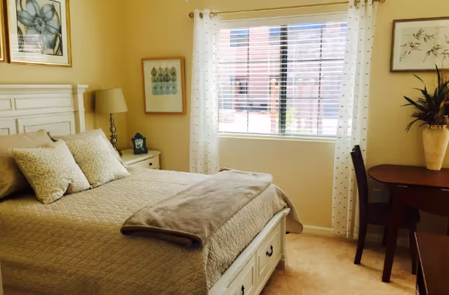 A cozy bedroom with a neatly made bed featuring beige quilted bedding and matching pillows. There is a white headboard and a nightstand with a lamp and a small decorative item. A window with white curtains lets in natural light, and a wooden table with a chair and a vase with plants is positioned near the window. The walls are painted a soft yellow and decorated with framed artwork.